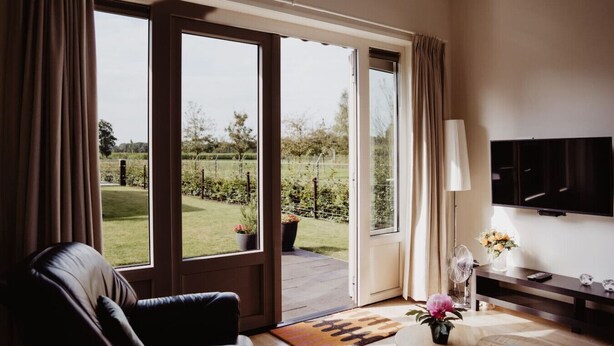 Un salon moderne avec un canapé en cuir, un téléviseur à écran plat, une petite table ornée d’un vase de fleurs et une grande porte-fenêtre coulissante donnant sur un jardin.