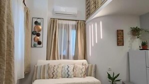 A small, cozy room with a bed, a window with curtains, a potted plant, and a white air conditioning unit. - Isidora Apartments (Zagora-Mouresi)