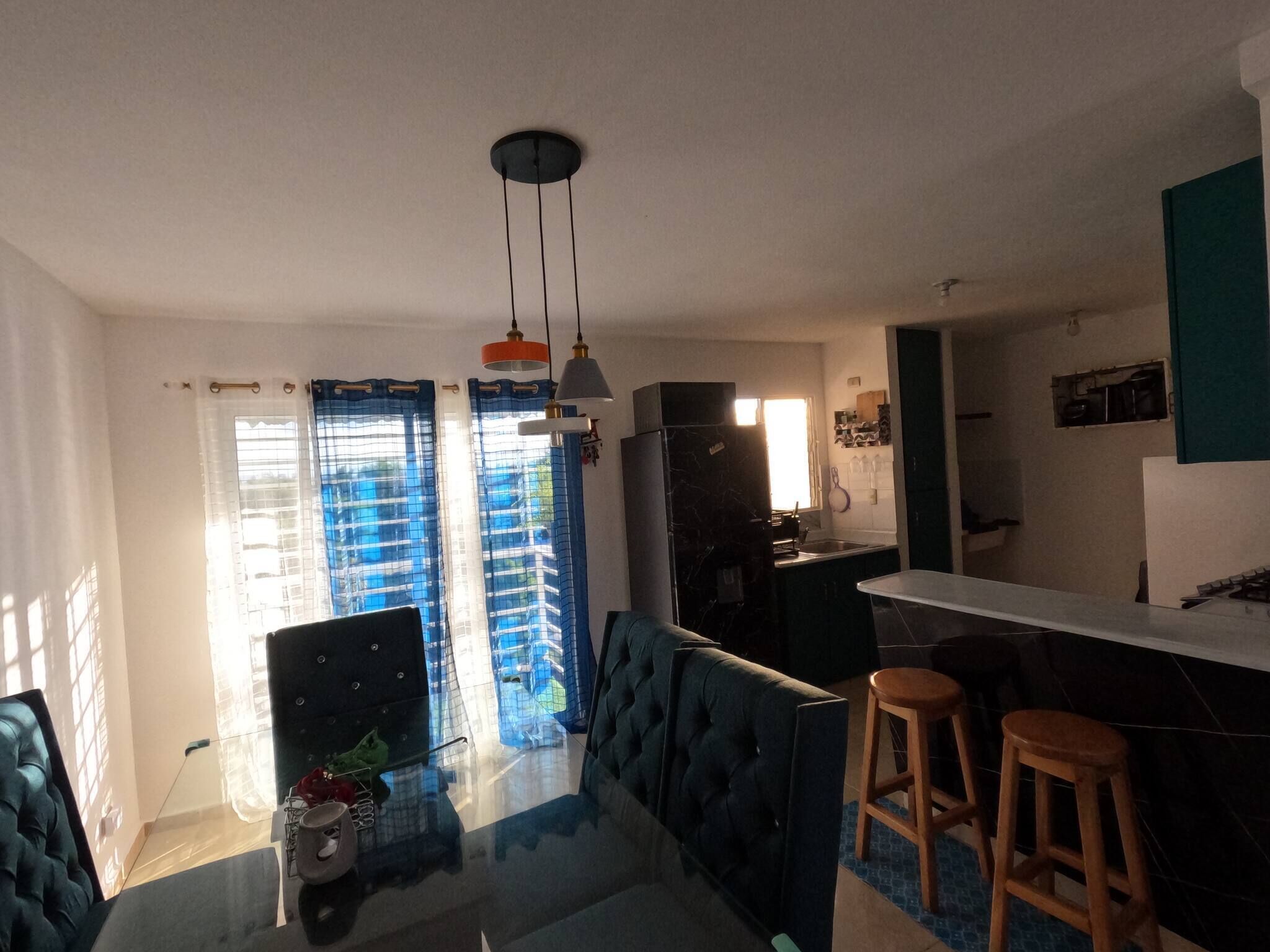 A modern kitchen with dark cabinets, a marble countertop, and a built-in oven. There is a dining area with a blue sofa and two wooden stools.