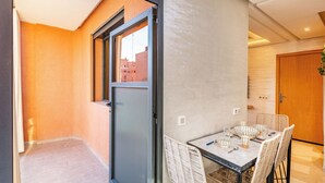 Un espace repas moderne avec une table dressée pour quatre personnes, entourée de chaises en osier, et une vue sur les bâtiments à travers une porte vitrée.