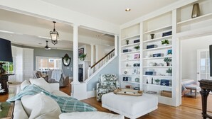 Un salon spacieux avec un canapé blanc, un fauteuil à motifs et une table basse en bois. Un escalier avec une rampe mène à une autre pièce.