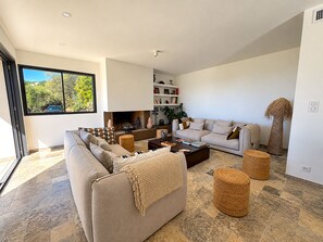 Ein modernes Wohnzimmer mit einer Couch, einem Couchtisch und einem Kamin. Ein Bücherregal und ein Fenster mit Blick auf Grünpflanzen sind ebenfalls vorhanden.