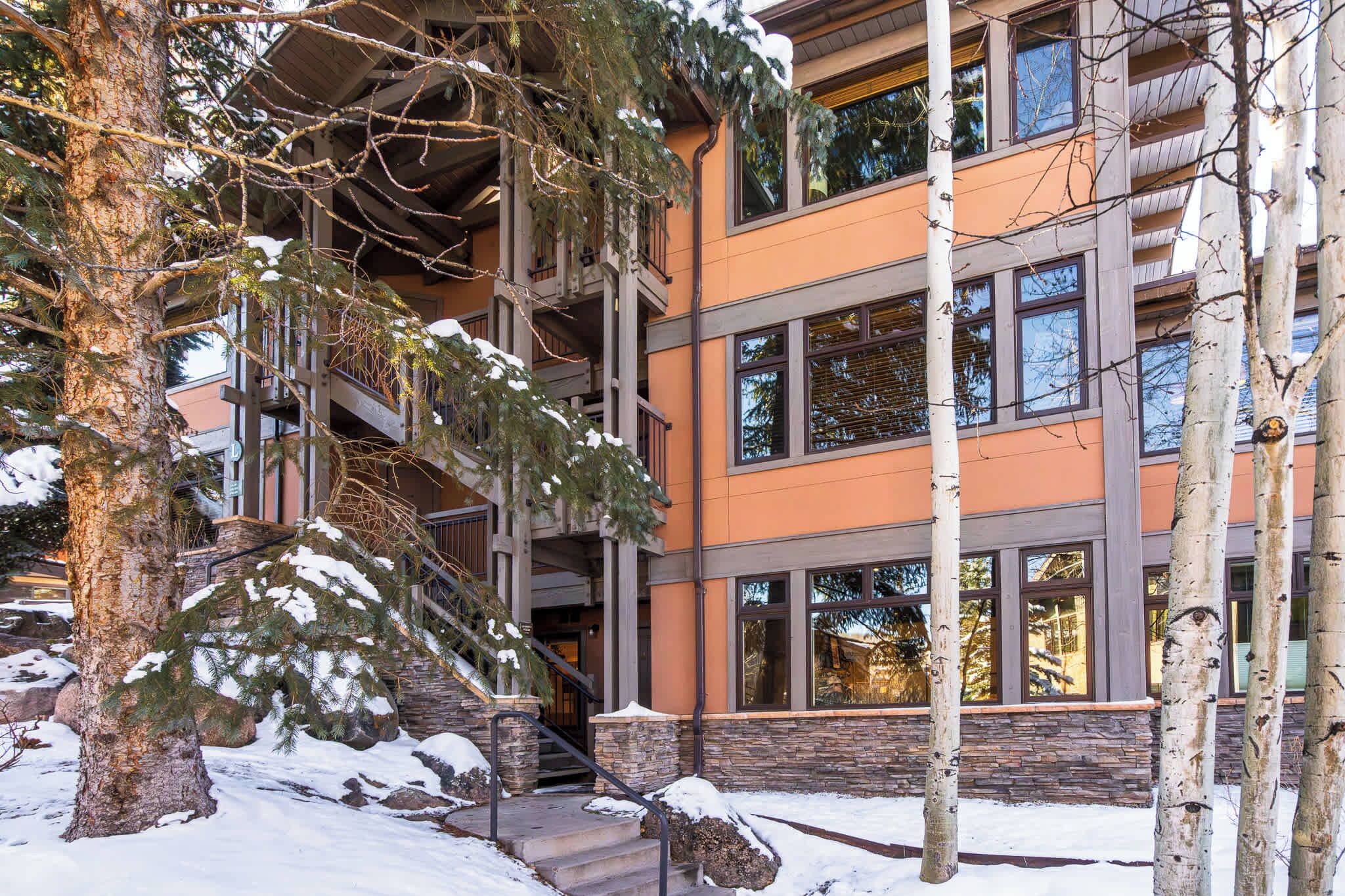 A multi-story building with a stone base and a wooden upper level, surrounded by snow-covered ground and trees. - New Ski-in Ski-out 1 BR with Resort Amenities (Snowmass Village)