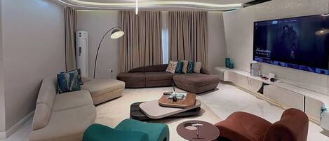 A modern living room with a large flat-screen TV, a brown sofa, a teal pouf, and a coffee table.