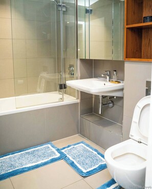 Een moderne badkamer met een glazen douchecabine, een witte badkuip, een wastafel met een spiegel erboven en een toilet. Op de vloer liggen blauwe en witte badmatten.