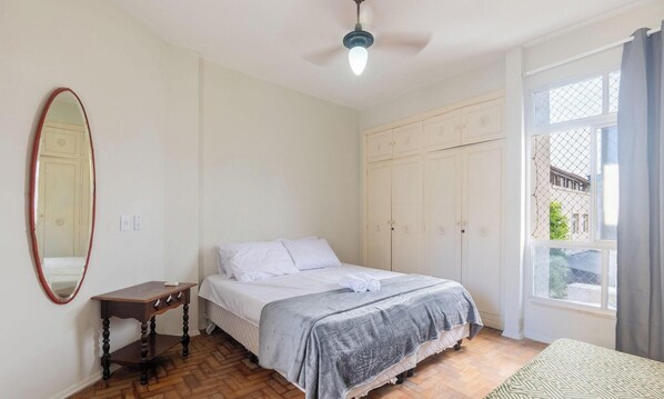 A bedroom with a bed, a wardrobe, a mirror, a small table, and a window with curtains. - 2-bedroom Apt Only 500m From the Beach Nos0201 (Guarapari)