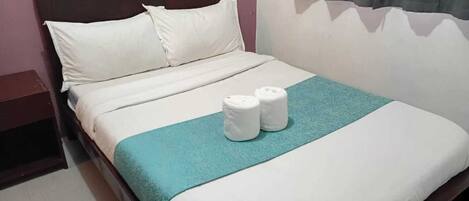 A neatly made bed with white linens and a teal accent, two rolled towels on the bed, and a wooden headboard.