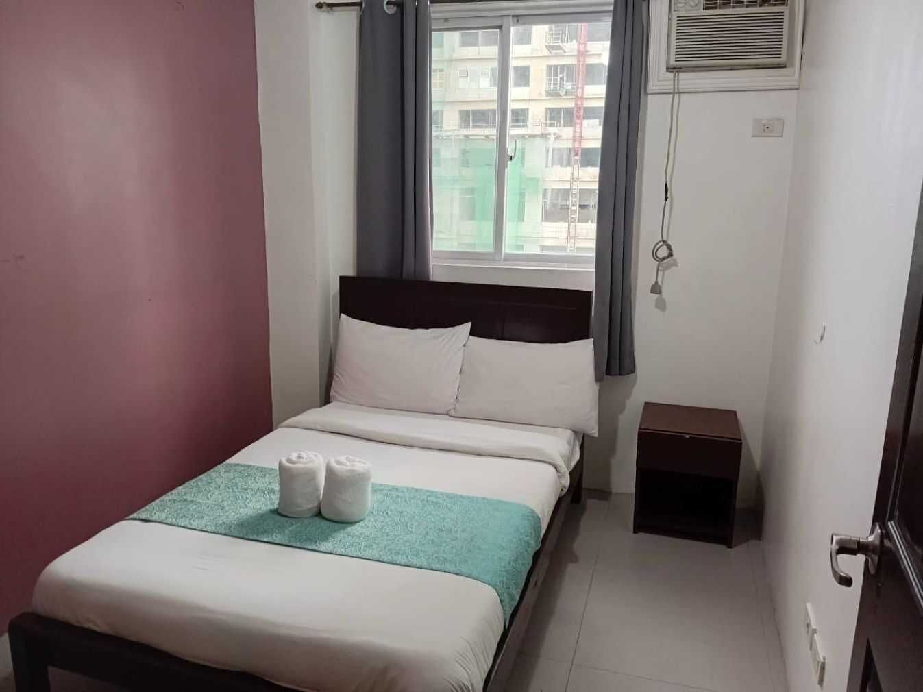 A neatly made bed with white bedding and a teal throw, a bedside table, and a window with curtains.
