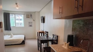 Une chambre d’hôtel compacte comprenant un lit, une petite table à manger avec des chaises, une kitchenette avec un évier et un téléviseur fixé au mur.