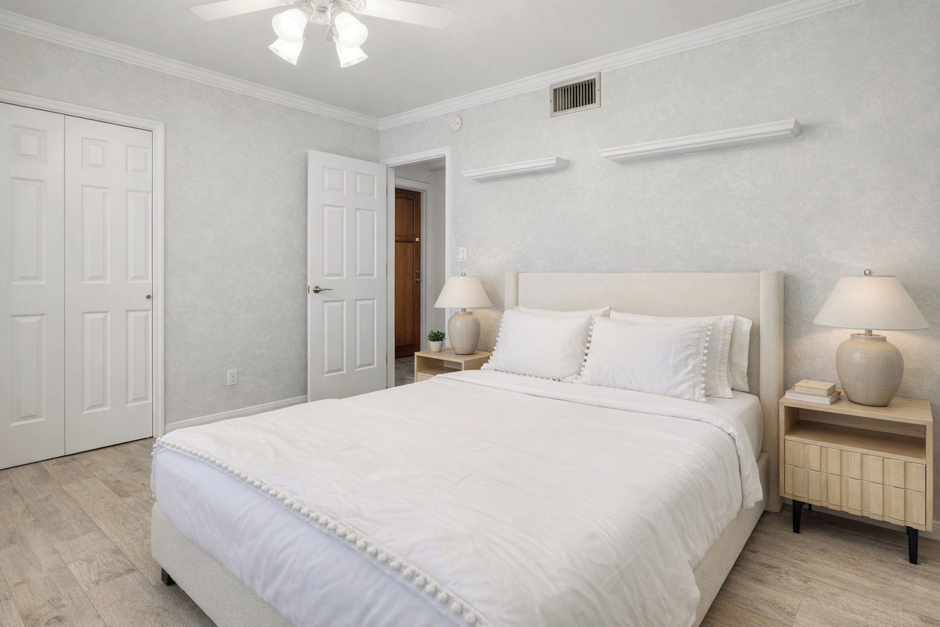 A bedroom with a large bed, two bedside tables with lamps, a ceiling fan, and a door leading to another room.