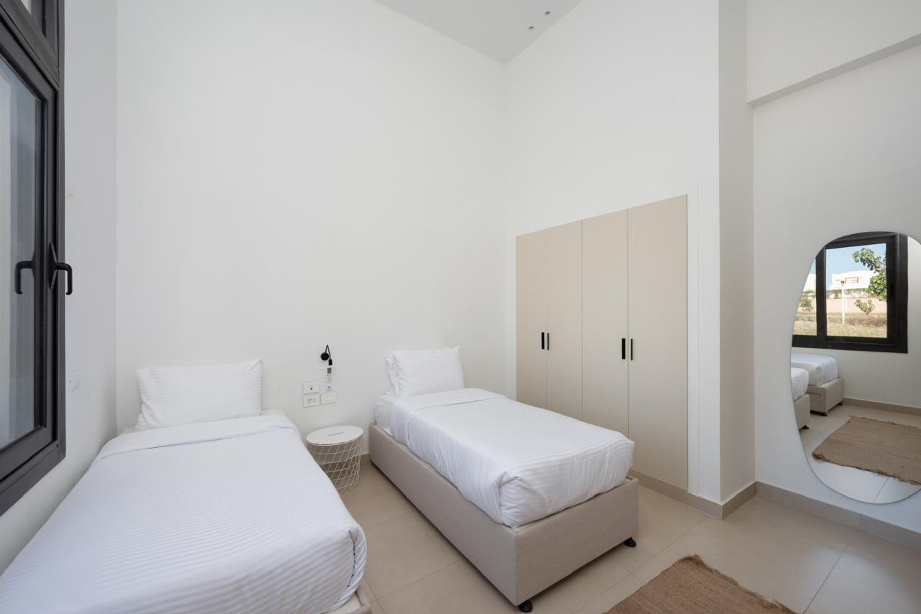Un dormitorio minimalista con dos camas, un armario, una mesita redonda y una ventana con vistas.