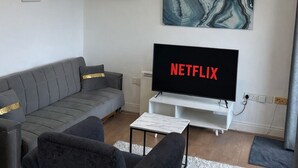 Een moderne woonkamer met een grijze bank, een zwarte fauteuil, een witte salontafel en een flatscreen-tv waarop 'NETFLIX' staat.