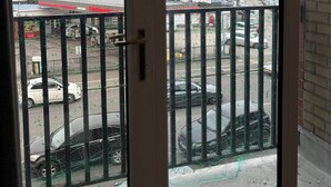 Uitzicht door een deur met een metalen hek, leidend naar een parkeerplaats met meerdere auto's en een gebouw op de achtergrond.