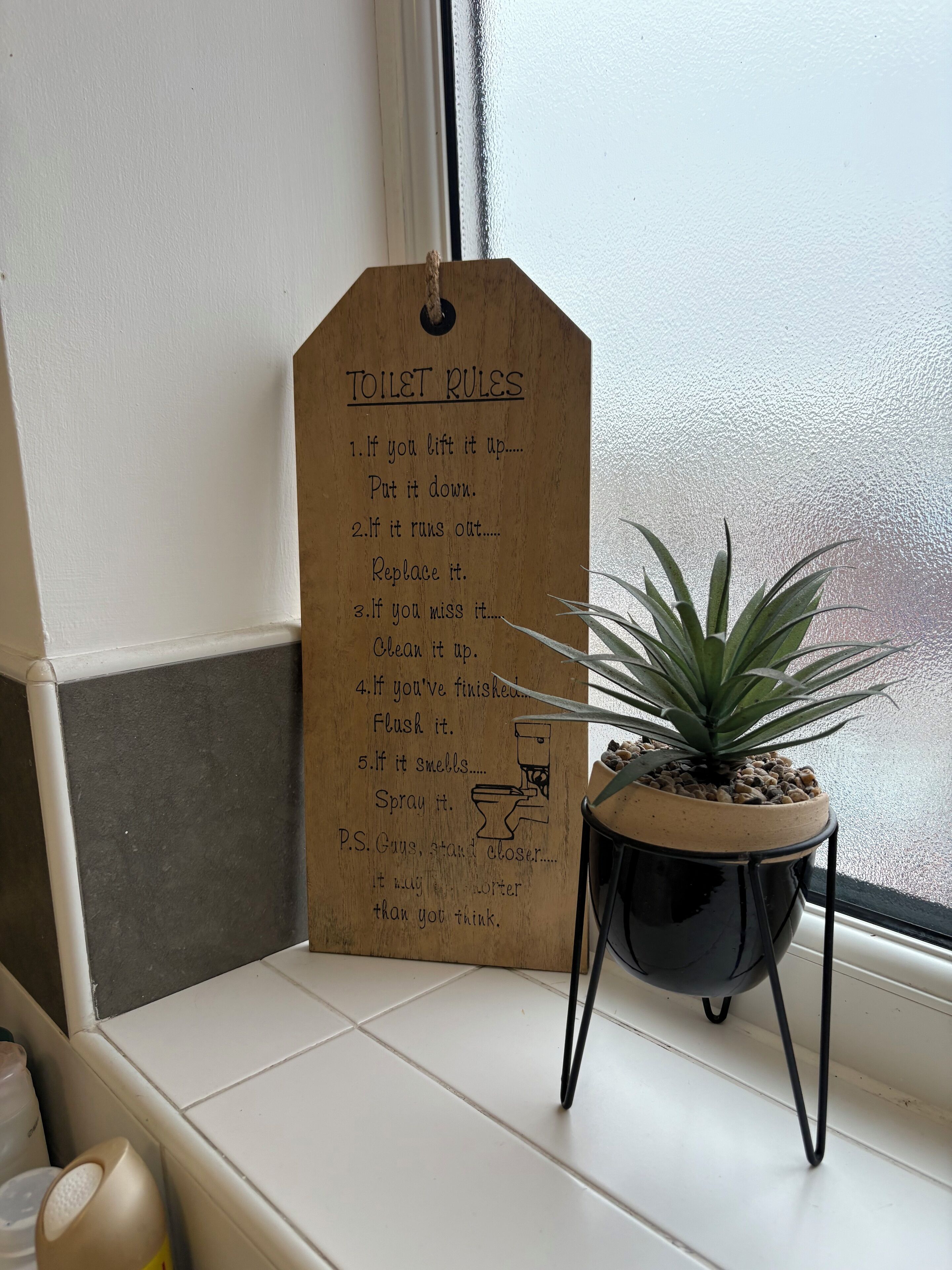 Un letrero de madera con normas del baño junto a una ventana con una planta en maceta en el alféizar.