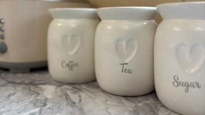 Drie keramieken potten met de tekst 'Koffie', 'Thee' en 'Suiker' op een marmeren aanrecht.