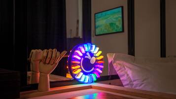 Une pièce comprenant une table sur laquelle est posée une tête de mannequin en bois, une horloge colorée et un lit recouvert de linge de lit blanc.