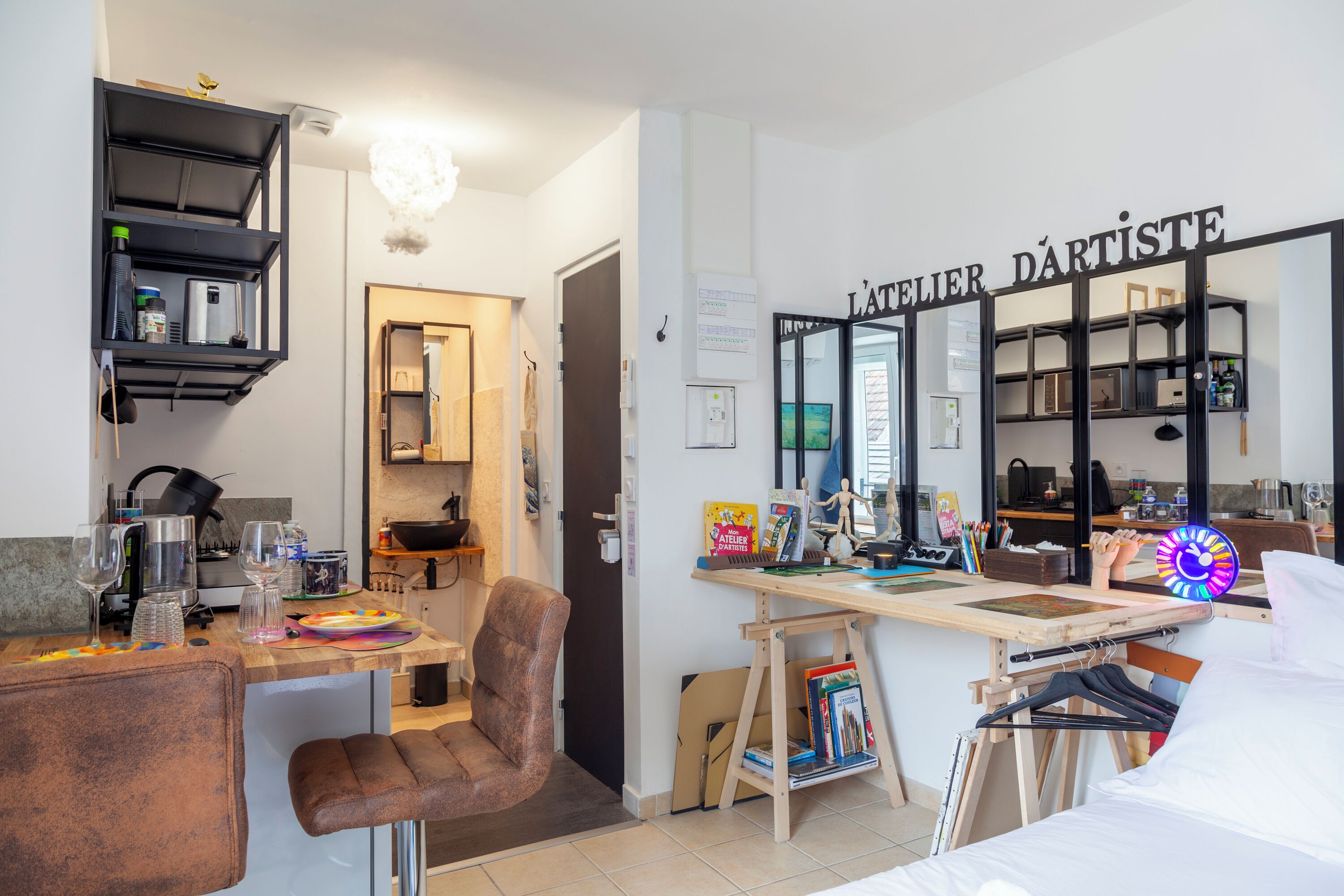 Une pièce moderne et bien éclairée, équipée d’un coin cuisine, d’un bureau avec un ordinateur et des livres, d’une chaise et d’un lit.