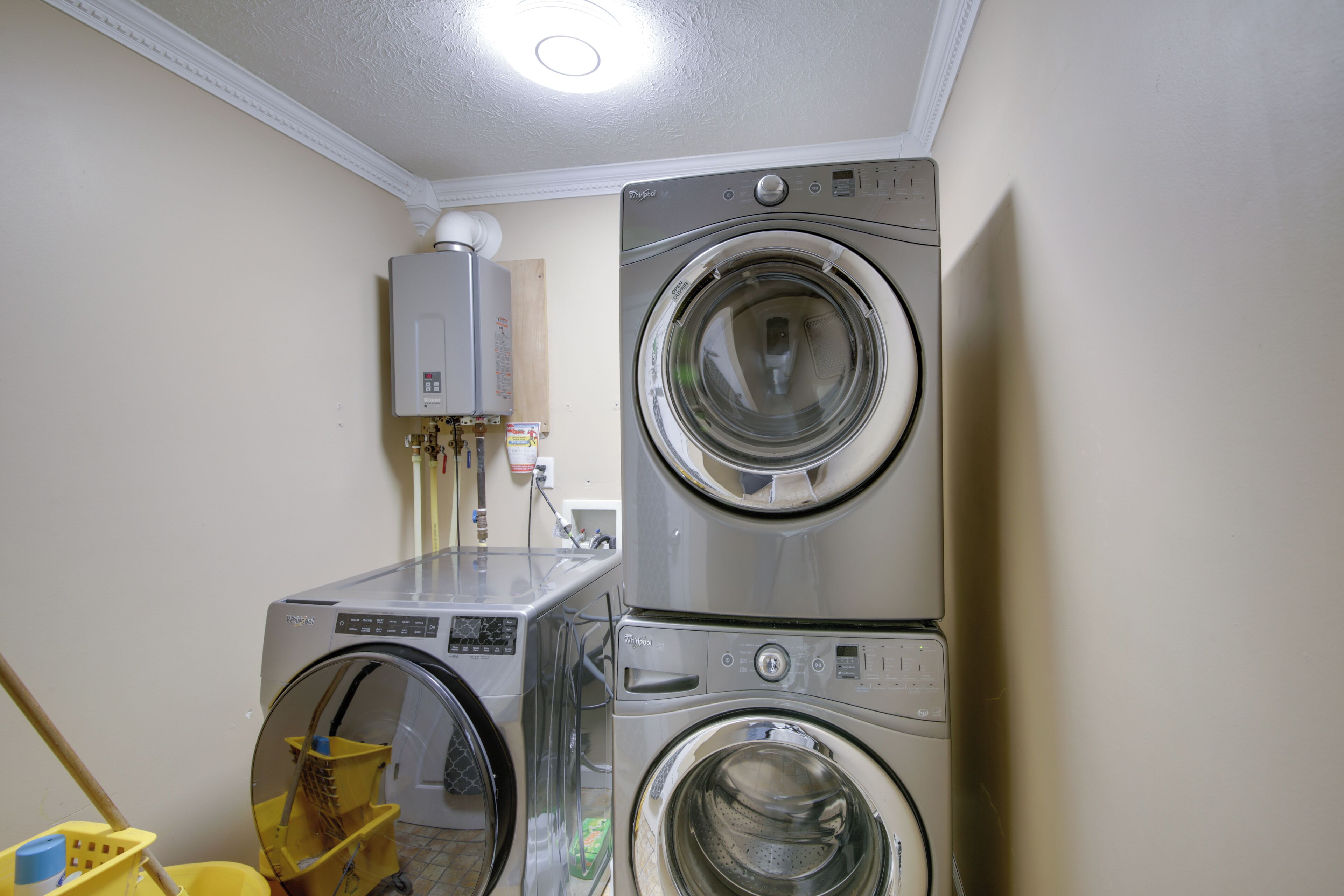 A laundry room with two stacked front-loading washing machines, a water heater, and a yellow mop bucket.