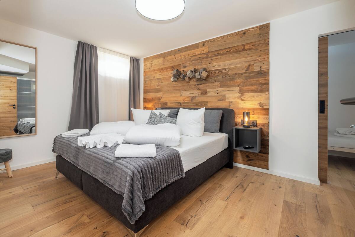 A modern bedroom with a wooden wall panel, a large bed with white and gray bedding, a bedside table with a lamp, and a wooden floor.