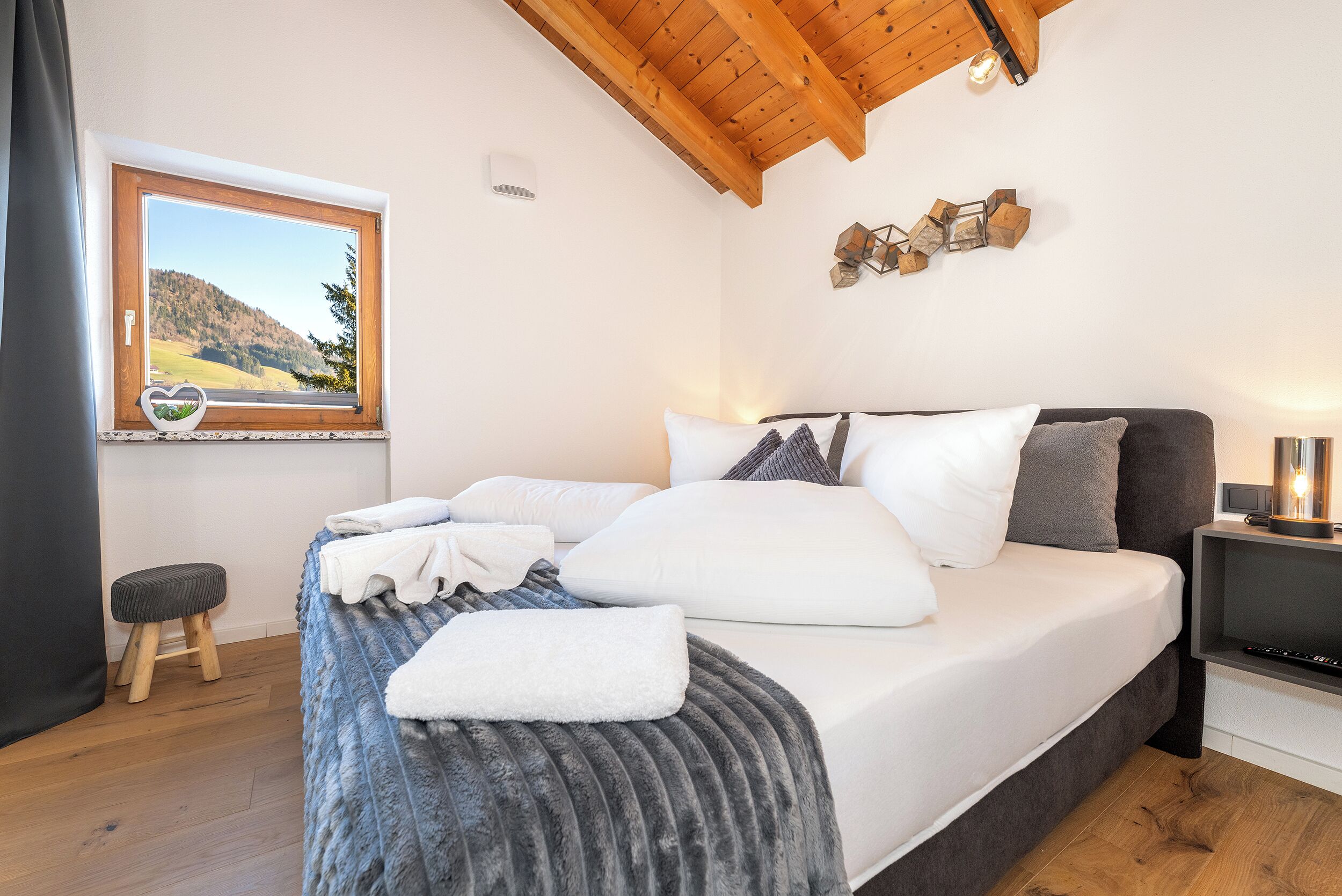 A bedroom with a bed, a window with a view of a landscape, a wooden ceiling, and a small stool.