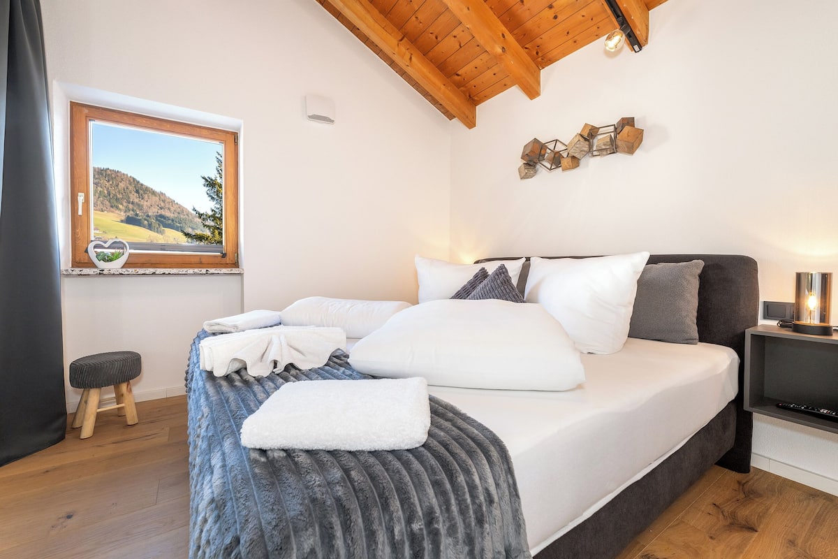 A bedroom with a bed, a window with a view of a landscape, a wooden ceiling, and a small stool.