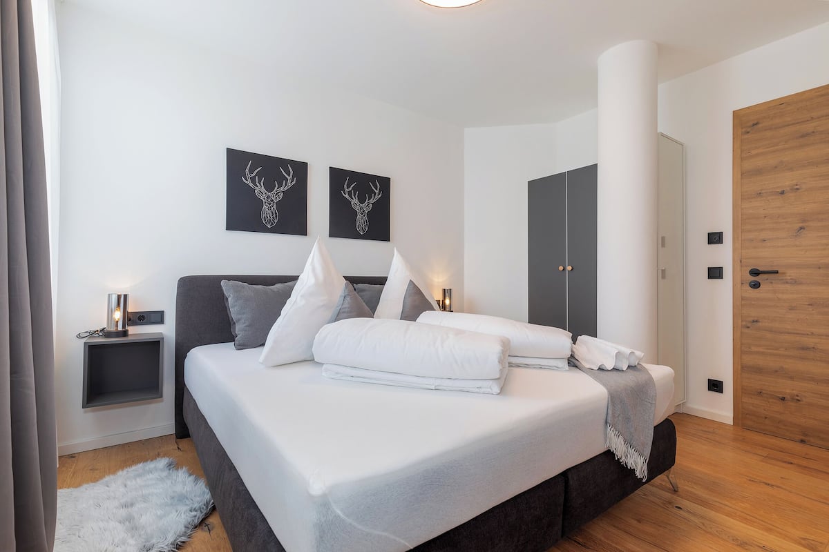 A modern bedroom with a large bed, two wall-mounted deer head art pieces, a bedside table with a lamp, and a wooden door.