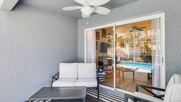 A covered patio area with a ceiling fan, a white sofa, a small table, and a glass door leading to a pool area.