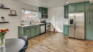 A kitchen with green cabinets, stainless steel appliances, a wooden floor, and a dining area with a table and chairs. - 3 Mi to Geha Field at Arrowhead: Easy Match Access (Independence)