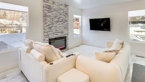 A modern living room with a fireplace, a flat-screen TV, and a large window with a view of a snowy landscape. - Newly Renovated, Modern Mtn Getaway: Pets Welcome! (Vail)