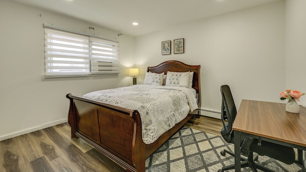 A bedroom with a wooden bed, a desk, and a chair. There is a window with blinds, a lamp, and two framed pictures on the wall. - Walk to Park & Public Transit: Jersey City Apt (Jersey City)