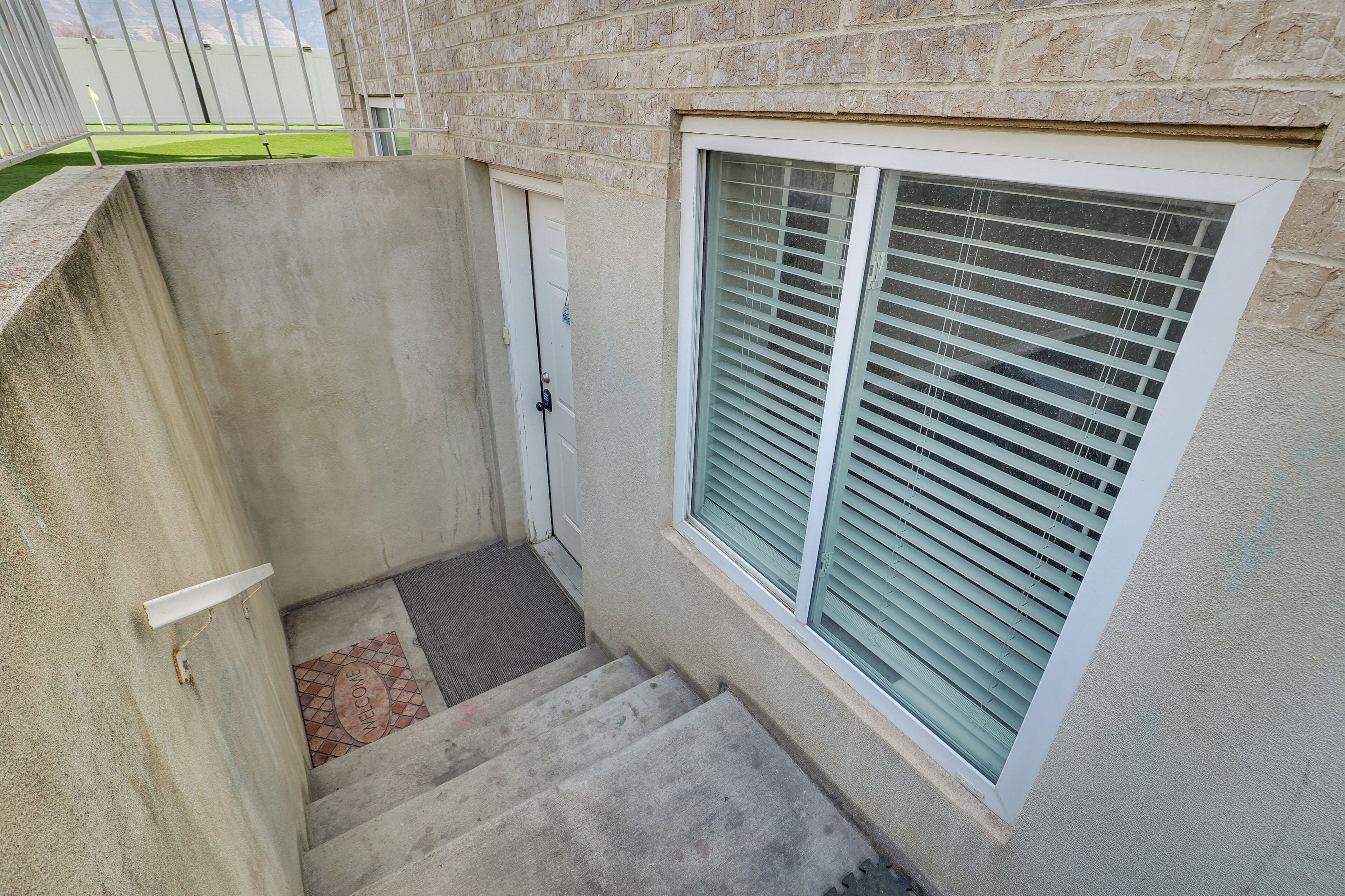 A concrete stairway leading to a door, a window with blinds, and a small patch of grass visible through a fence.