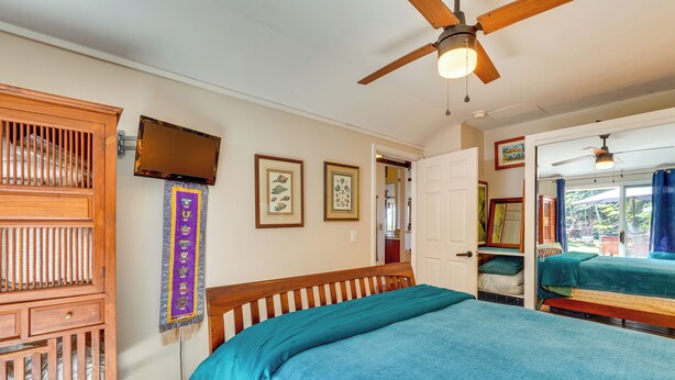 A bedroom with a large bed, a wooden cabinet, a ceiling fan, and a television on the wall. - Lush Outdoor Space & Epic Views! Kailua-kona Home (Kailua-Kona)