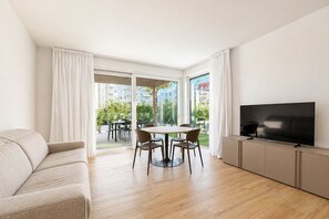 A modern living room with a sofa, a dining table with chairs, a flat-screen TV, and a sliding glass door leading to an outdoor area. - Aurora Sand 1 - Carraro Immobiliare Jesolo (Jesolo)
