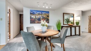 Et spiseområde med et rundt træbord, fire grå polstrede stole, en hvid vase med blomster, et stort landskabsmaleri, et spejl og et konsolbord med en lampe og dekorative genstande.