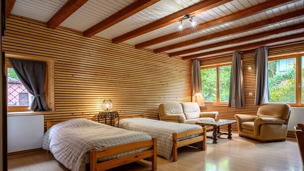 A room with two wooden beds, a sofa, a chair, a table, and large windows with curtains. - Gîte Baumann Rombach le Franc – Cozy House with Terrace (Rombach le Franc)