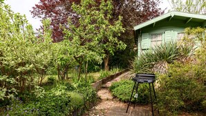 A wooden deck with a grill, surrounded by lush greenery and a small building with green shutters. - Gîte Baumann Rombach le Franc – Cozy House with Terrace (Rombach le Franc)