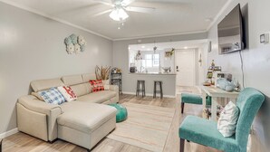 Una sala de estar con un sofá seccional color beige, sillones color turquesa, un ventilador de techo y una zona de cocina al fondo.