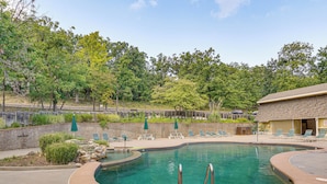 A swimming pool with a curved design, surrounded by lounge chairs and umbrellas, set against a backdrop of trees and a stone wall. - Private Dock at Ozarks Home Next to Margaritaville (Osage Beach)