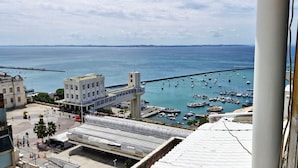 Un paysage urbain côtier avec un port de plaisance rempli de bateaux, un imposant bâtiment blanc surmonté d’une tour, et un pont piétonnier enjambant l’eau.