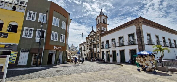 A cobblestone street with historic buildings, a church tower, and a market stall. - COPSE0103 - Edifício Themis (Salvador)