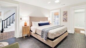 A bedroom with a large bed, a nightstand with a lamp, a ceiling fan, and a view of the outside through a window. - Hidden Garden Retreat in the Heart of Downtown Savannah (savannah)