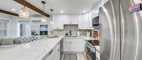A modern kitchen with white cabinets, a large stainless steel refrigerator, pendant lights, and a herringbone tile backsplash.