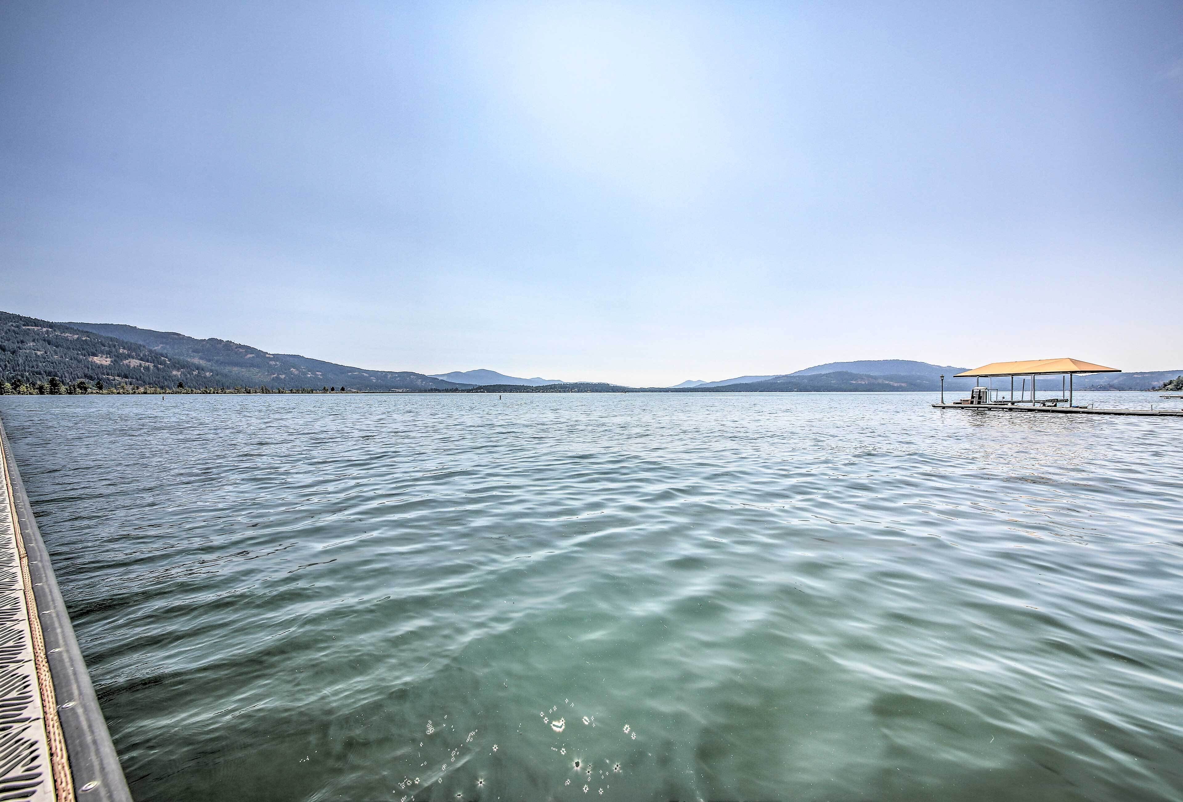 A calm lake with a wooden dock extending into the water, a small pavilion with a canopy, and distant mountains under a clear sky.