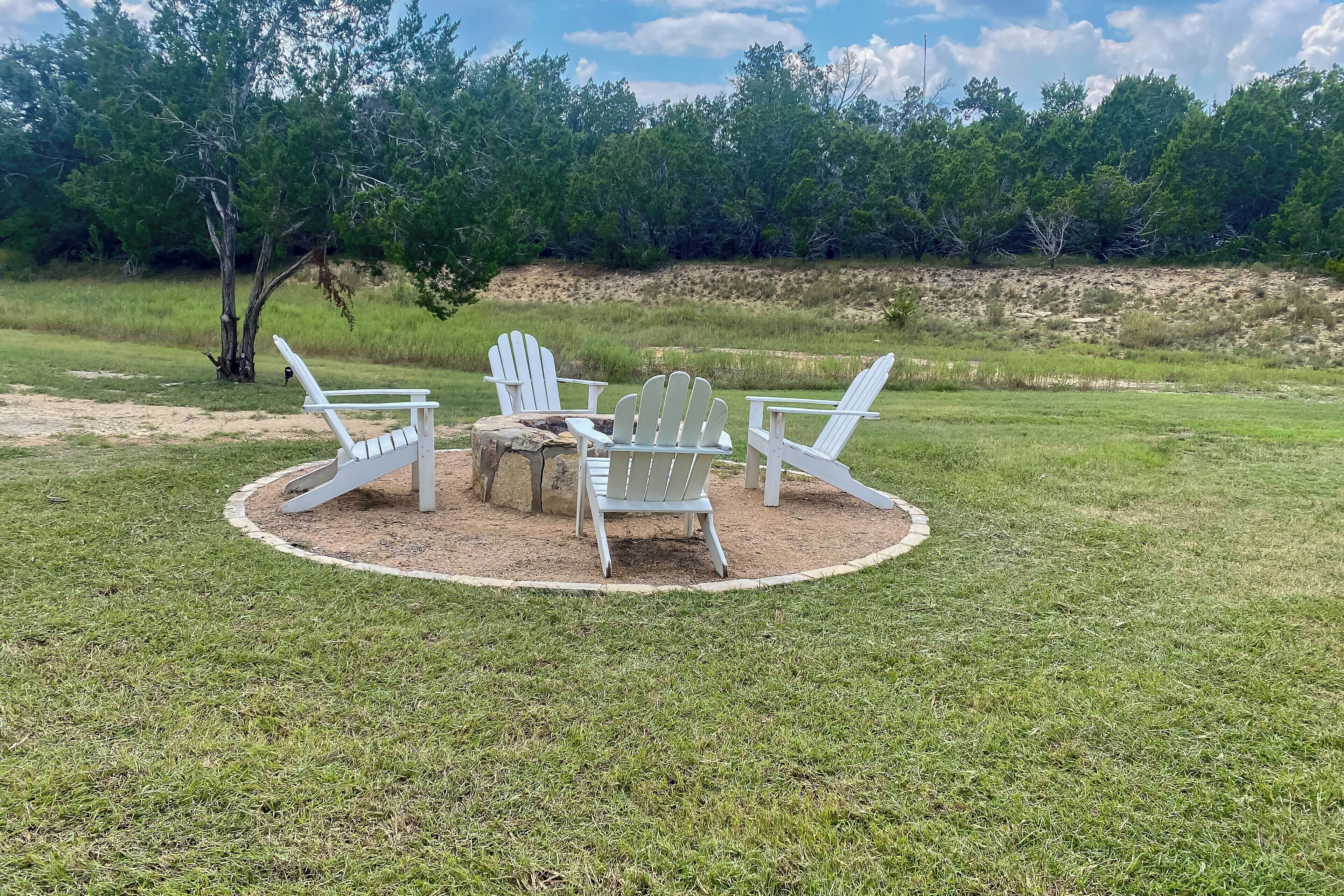 A circular outdoor seating area with Adirondack chairs and a small table, surrounded by a grassy field and trees.
