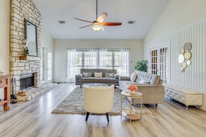 A spacious living room with a fireplace, a ceiling fan, and a large window with curtains. - Hill Country Escape: Family Home w/ Outdoor Oasis (Dripping Springs)