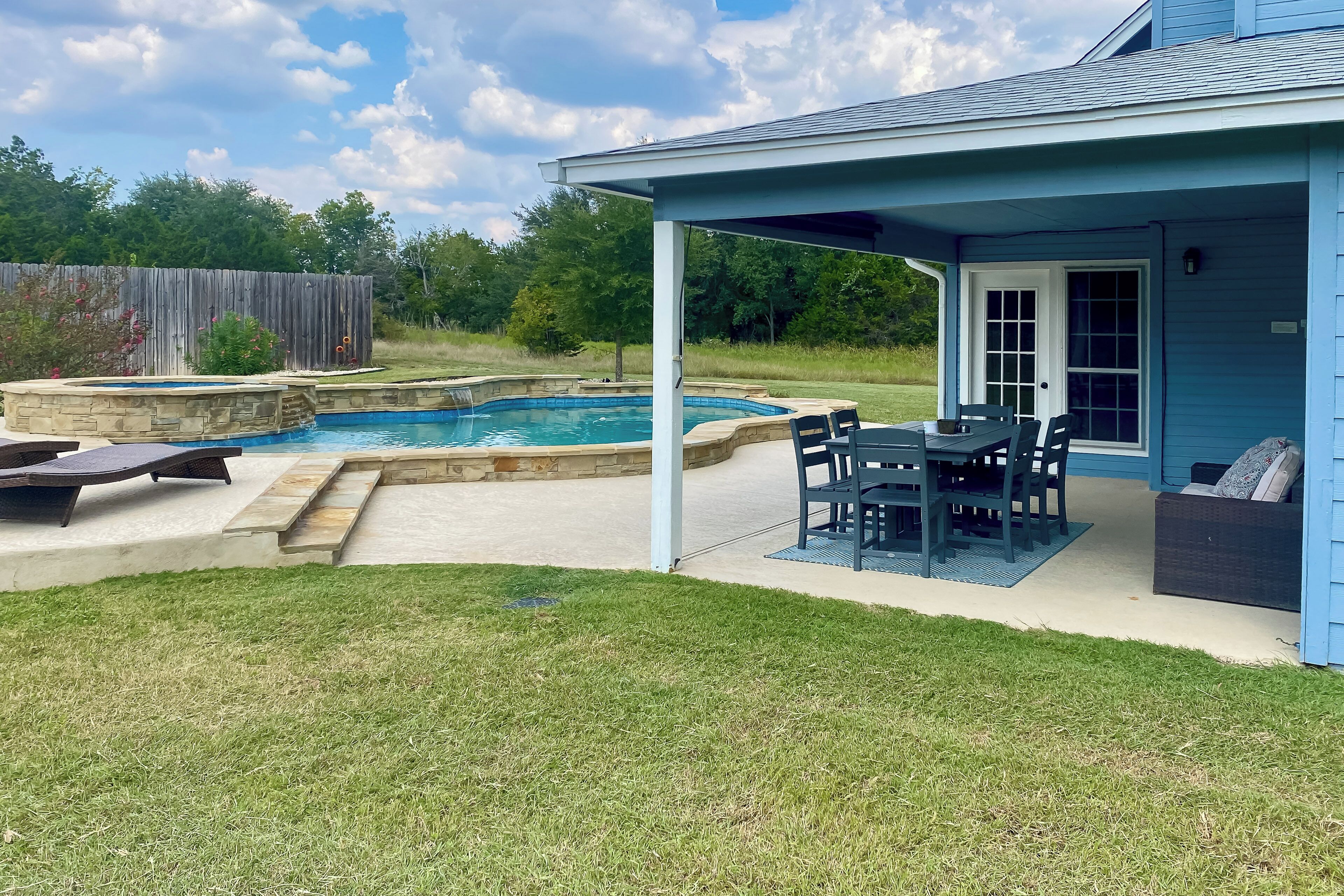 A backyard with a swimming pool, a covered patio with outdoor furniture, and a wooden fence.