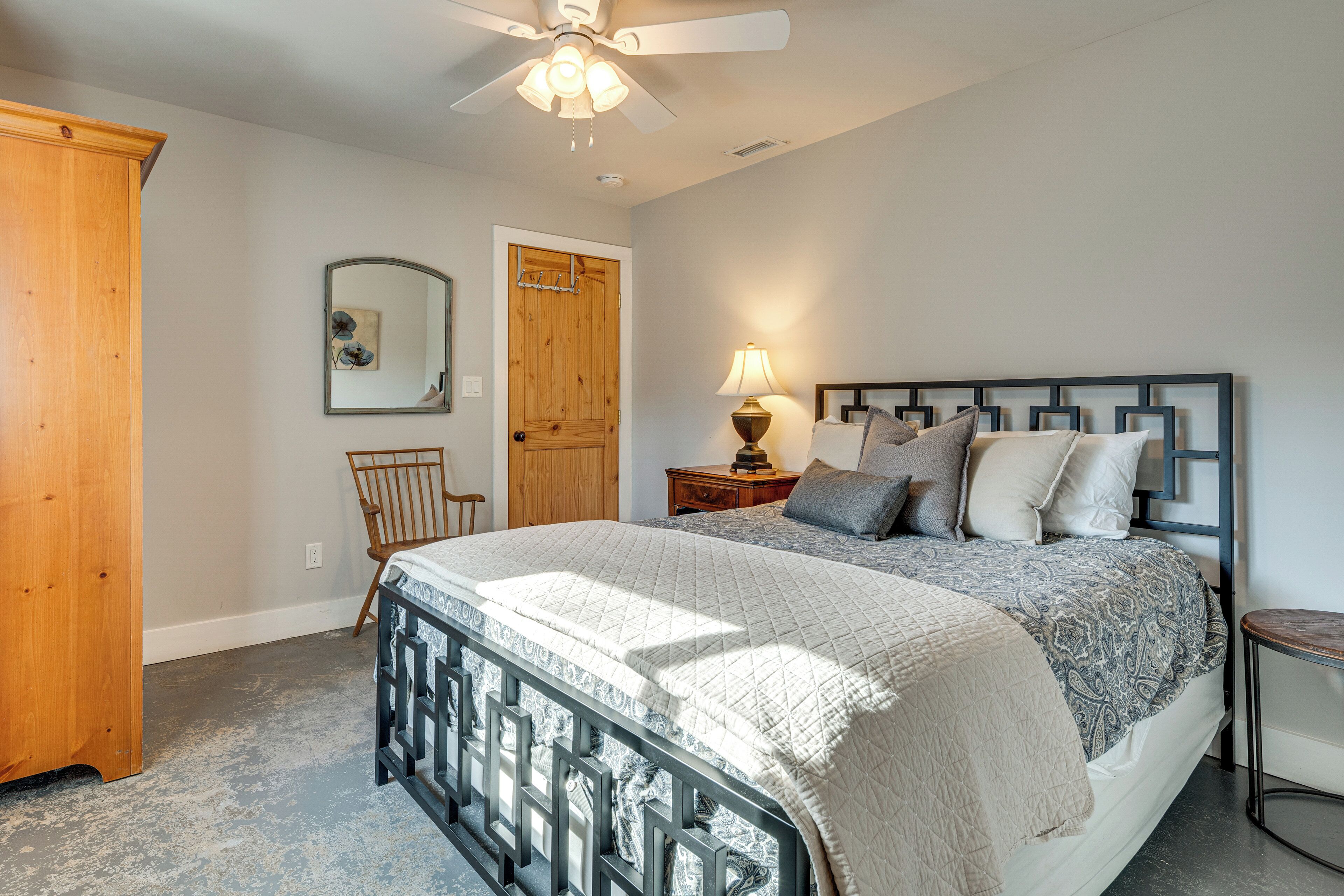 A bedroom with a bed, a wooden wardrobe, a chair, a nightstand with a lamp, and a ceiling fan.