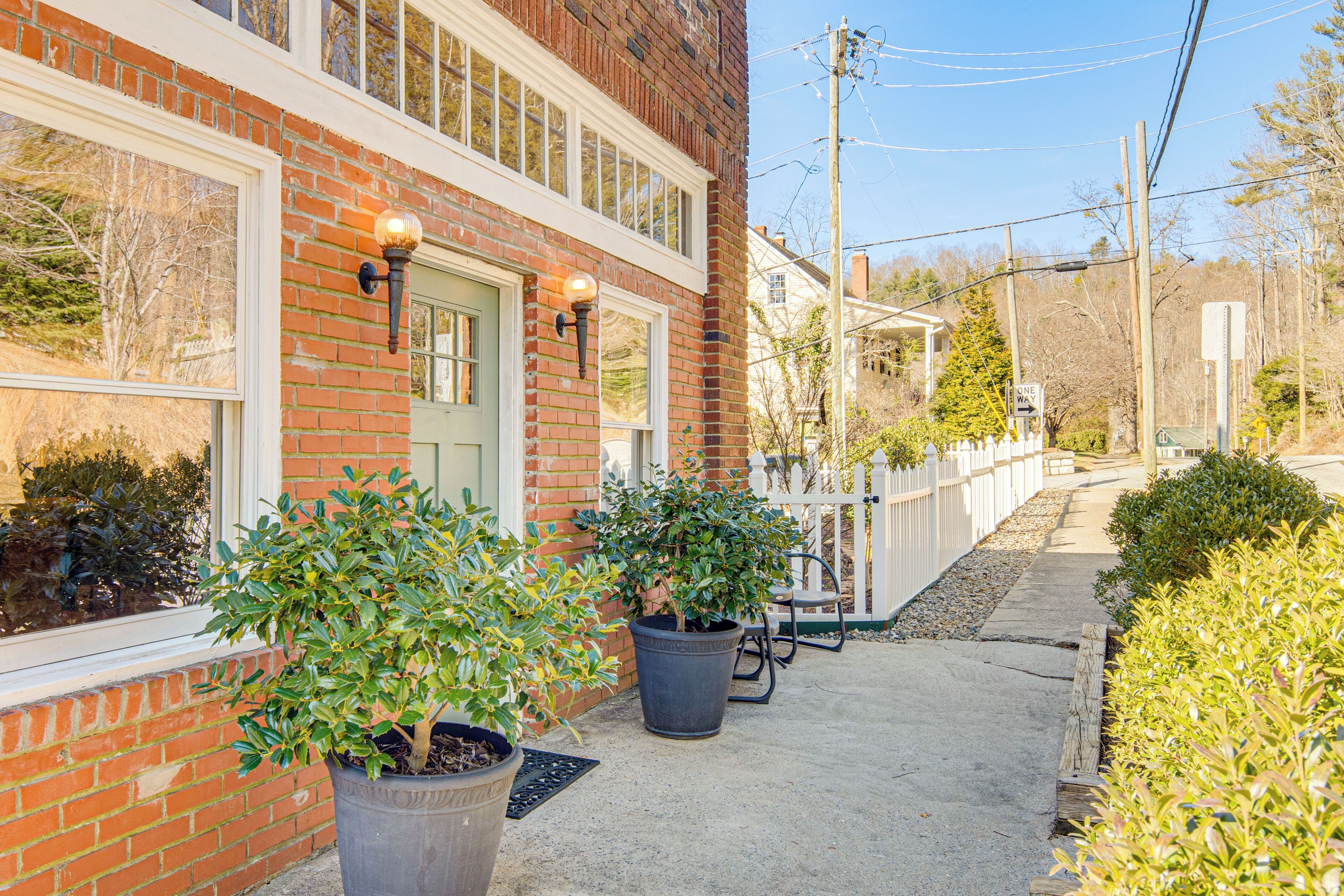 A brick building with a green door and windows, potted plants, a white fence, and a 'ONE WAY' sign.