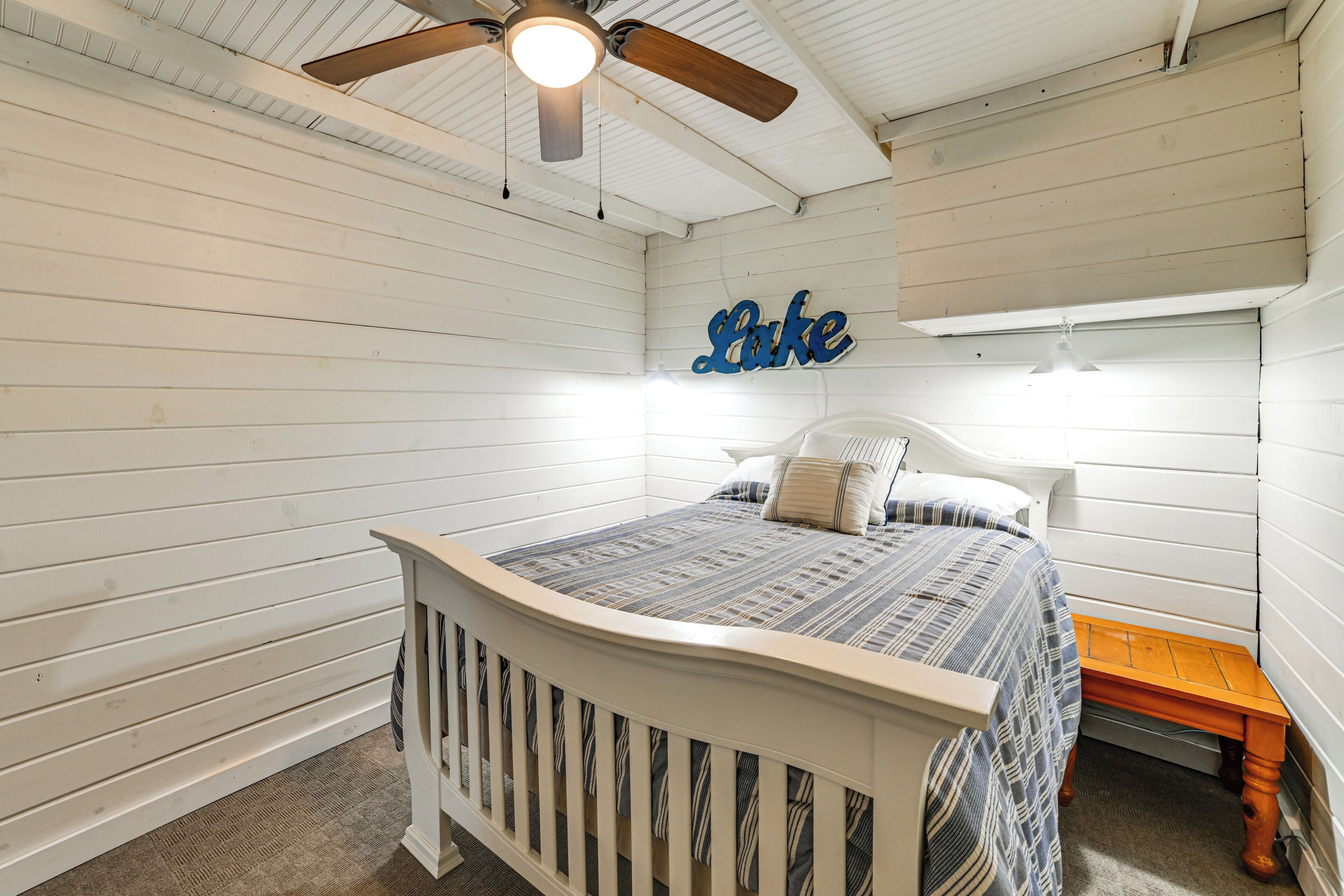 A bedroom with a white bed, a wooden nightstand, a ceiling fan, and a wall-mounted sign.