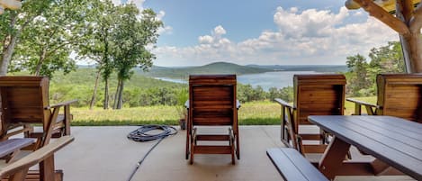 A covered outdoor seating area with wooden benches and picnic tables, overlooking a scenic landscape with trees and a body of water.
